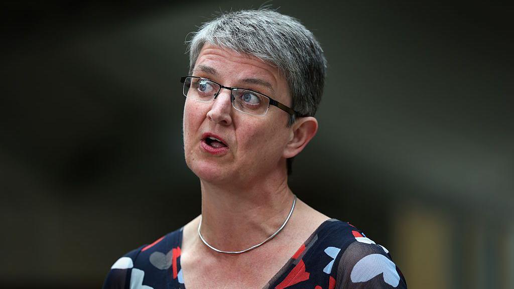 Maggie Chapman has short black/grey hair. She is wearing glasses and a blue floral top