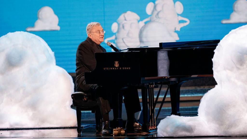 Musician Randy Newman plays a piano on stage with cut out clouds around him
