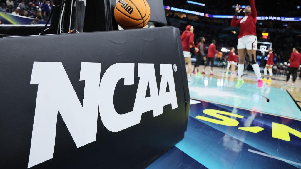 The NCAA logo pictured in front of a basketball court with players practicing in the background