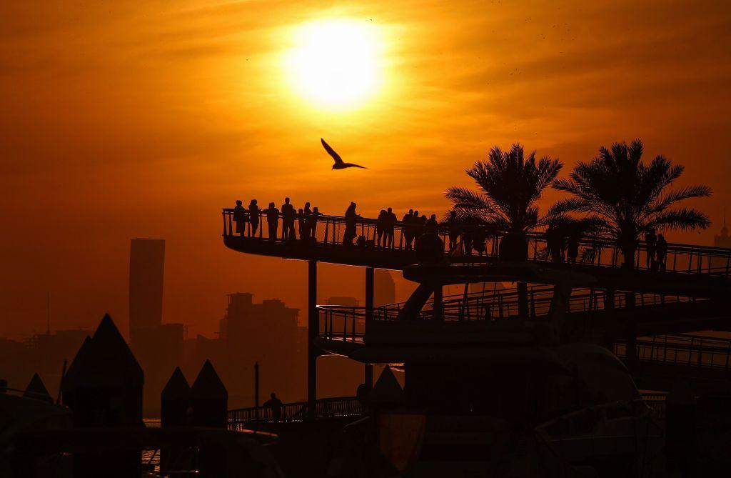 A group of people gather on a raised walkway in Dubai Creek Harbour at sunset. There is a large bird flying above them. There are skyscrapers in the distance and the orange of the sunset puts everything else into silhouette.
