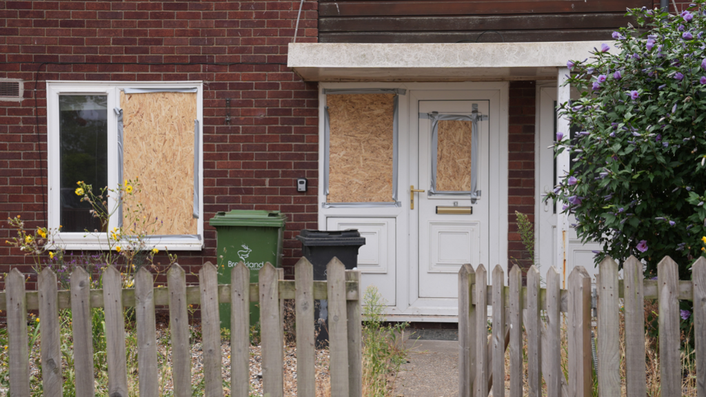 Image shows a boarded up house with bins outside and a weather-beaten picket fence in the foreground