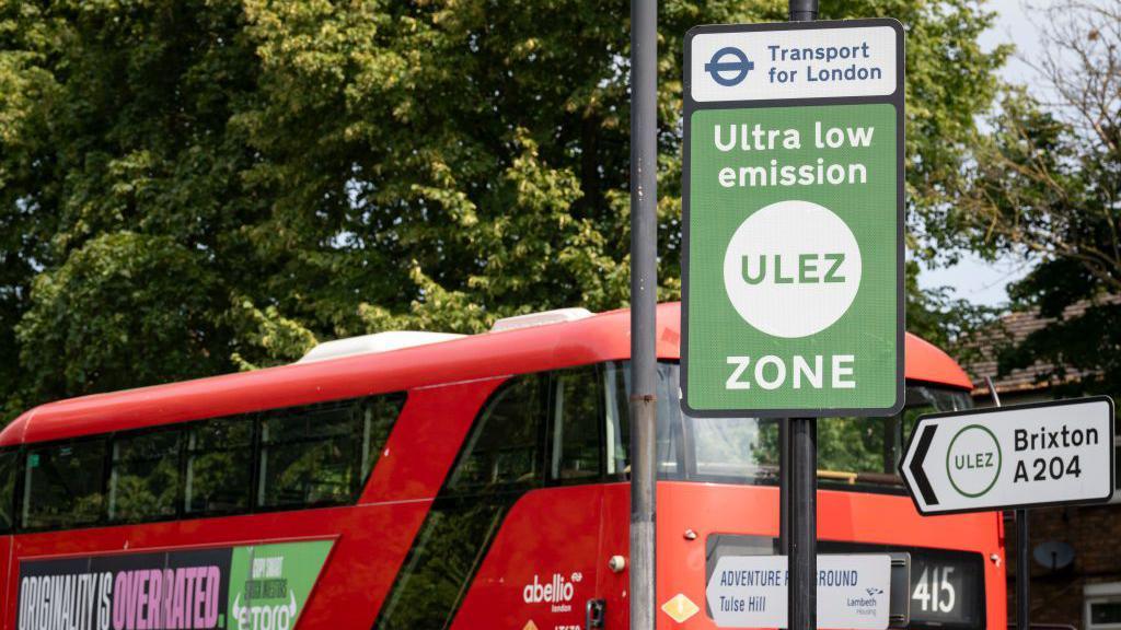 File image of a green and white Ulez road sign, with a red double decker bus passing behind it on a tree-lined road