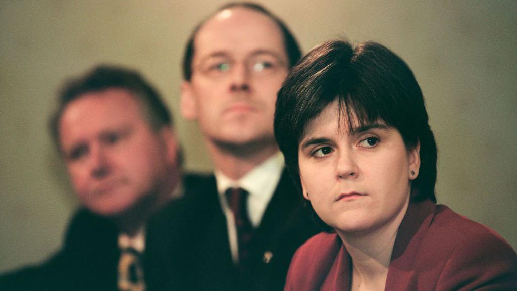 Nicola Sturgeon, on the right of the picture, at the launch of the SNP's 1999 manifesto. She is wearing a maroon jacket and is looking to her left.