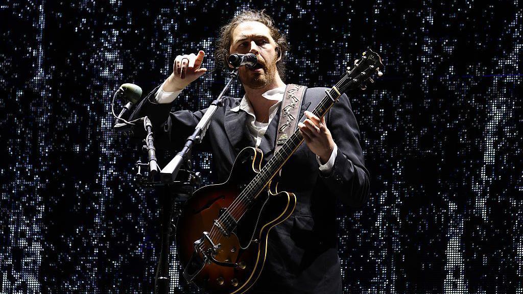Hozier, a man with a beard and long brown hair tied back, points with his right hand as his hand rests on the fingerboard of his electric guitar.