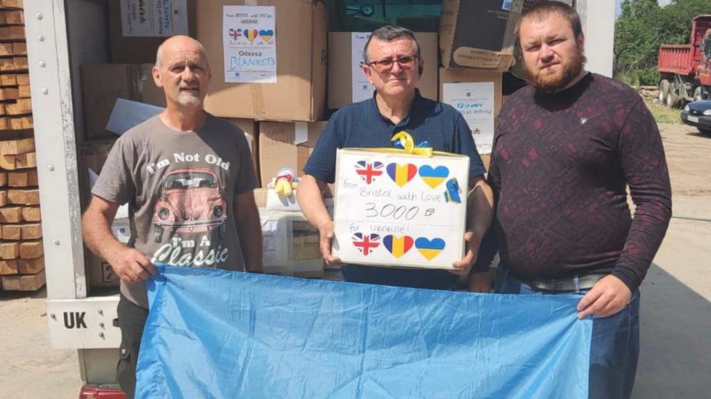 Three men standing in front of a lorry with boxes in the back. They are holding a Ukrianian flag