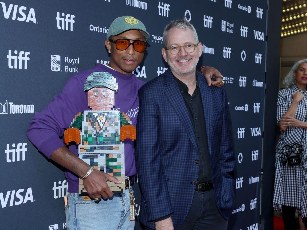 Pharrell Williams and Morgan Neville at the world premiere of Piece By Piece at the Toronto Film Festival. Williams is standing on the left wearing light blue jeans and a purple top and is holding a Lego model of himself. Neville has short grey hair and glasses and is wearing a black and navy checked suit.