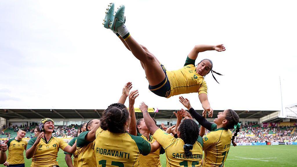 A Brazilian rugby player in a yellow shirt being thrown in the air by her teammates.