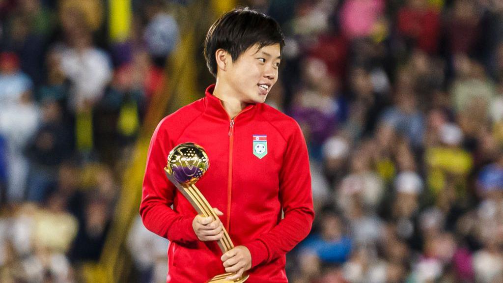 Choe Il-son, wearing his country's red tracksuit, smiles as she holds the Golden Ball trophy