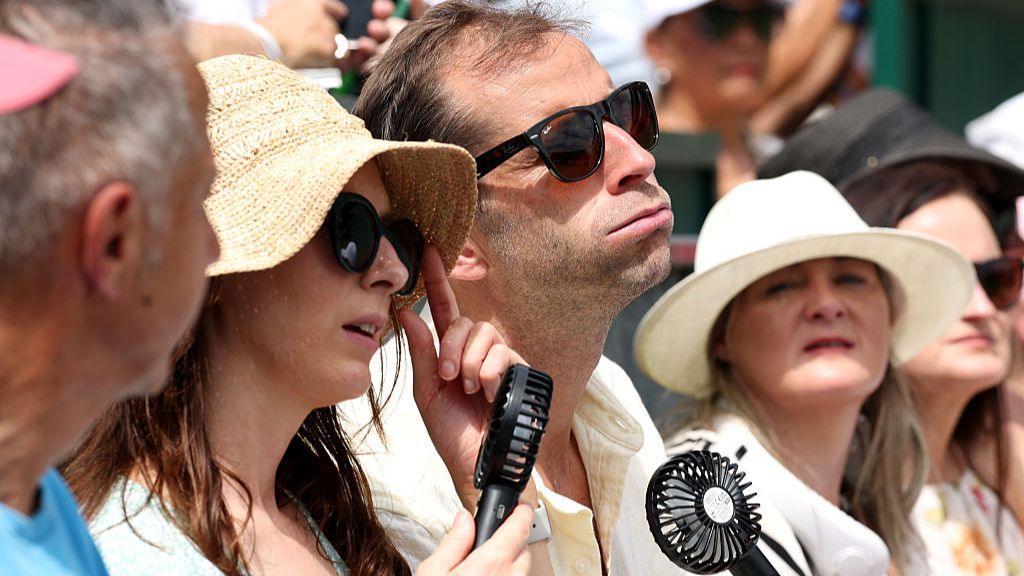 A row of people sat. The picture shows only their heads. A man in the middle looks like he is puffing in the heat, as he uses a hand fan and wears sun glasses. The women on either side wear wide straw hats, one wears sunglasses and also has a fan.