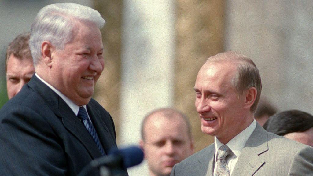 A younger looking Vladimir Putin stands in front of Boris Yeltsin, both in suits and laughing