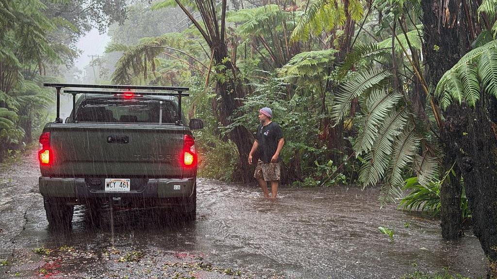 A car stops on a rural road as it floods in Volcano Village on Hawaii Island. A man stands next to the car as rain pelts down on a leaf-lined road