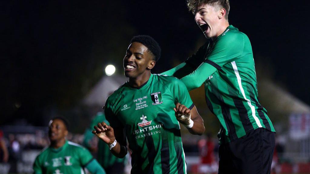 Kyrell Lisbie celebrates with teammate at a previous FA Cup game. He is wearing a green football shirt with black stripes.