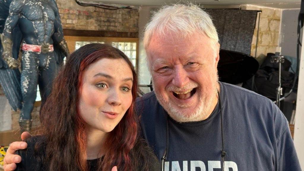 Grace Cope stands with Chris Kimsey. She wears a black T-shirt with red dyed streaks in her hair and is smiling. He wears a navy T-shirt and is laughing.