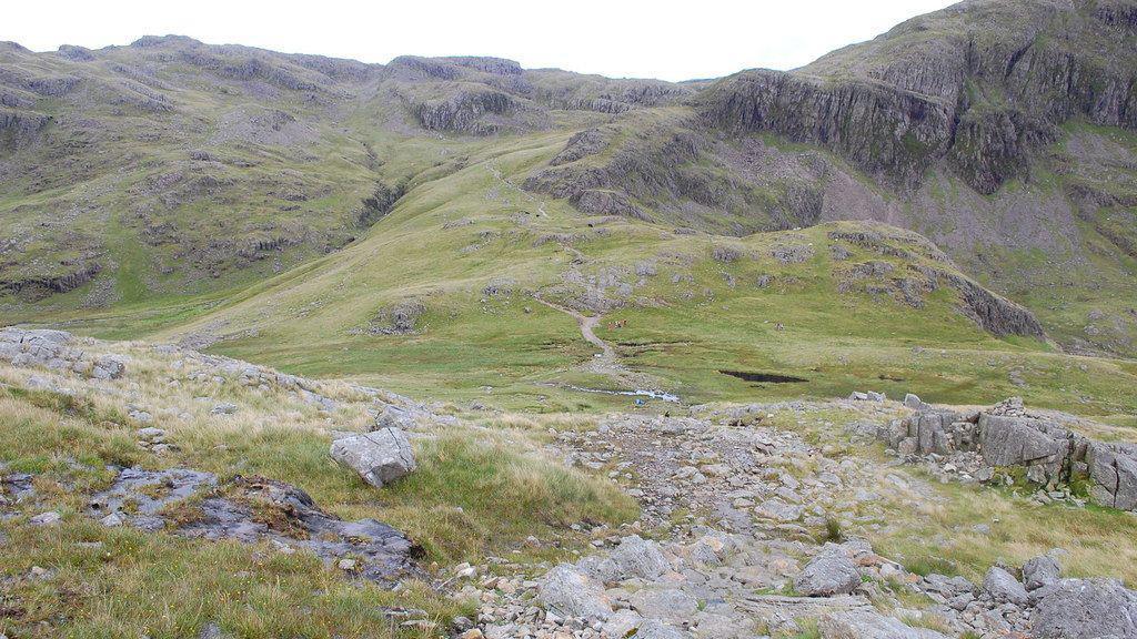Looking back down on Sty Head while ascending Great Gable by the Breast Route. The path to Sprinkling Tarn is ahead centre. There is a rocky path leading up to a crag which is covered in green grass.