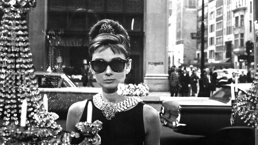 A black and white image of Audrey Hepburn taken from Breakfast at Tiffany's. Her character is wearing sunglasses and a sleeveless black shift dress with a thick necklace of pearls, and a tiara with her hair in a bun. She stands on the street looking into the window of a Tiffany's store displaying diamond jewellery and chandeliers