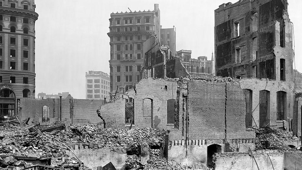 A black-and-white photograph of San Francisco streets in ruins after the earthquake. Several buildings have collapsed and the street is filled with debris