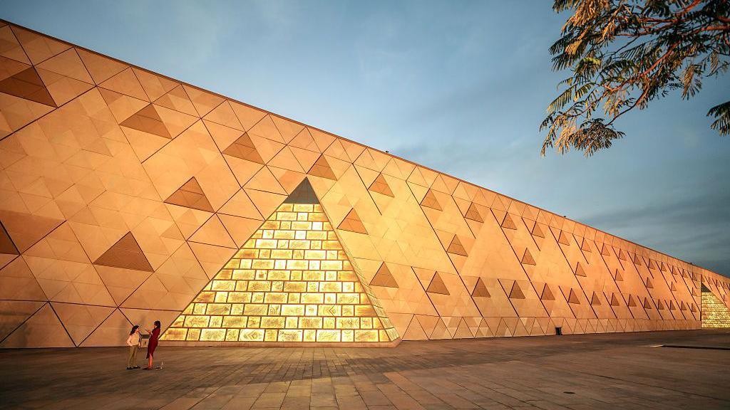 A side view of the museum at night, with a man standard next to one ofd the walls that has a brick-like pattern similar to a pyramid. Bright lights can be seen in the night sky. 