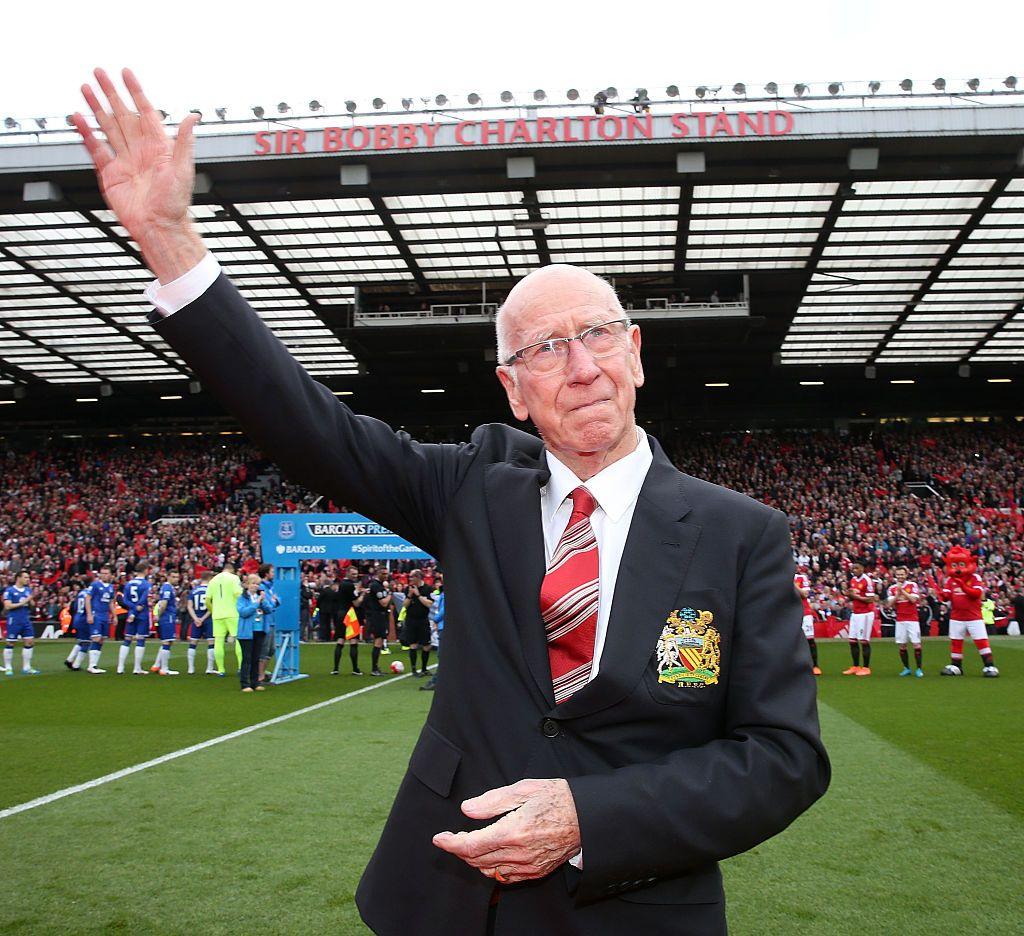 Sir Bobby Charlton, wearing a black blazer, white shirt and red striped tie, stands on the pitch at Old Trafford waving to crowds. He is attending the unveiling of a stand renamed in his honour, which can be seen packed with fans in the background