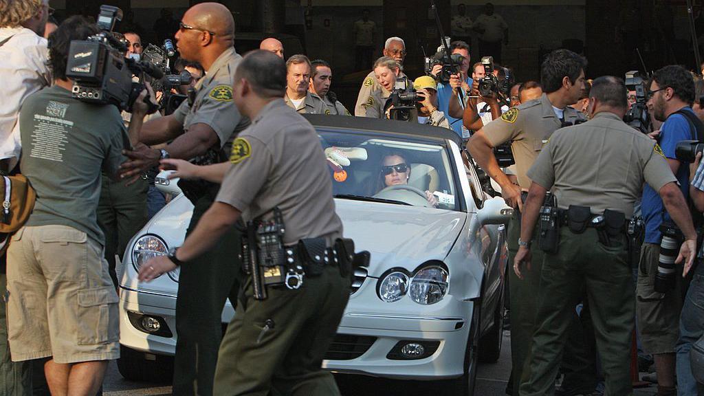 Britney Spears is seen driving a white sports car as people and media cameras surround her. Police officers are helping clear a path