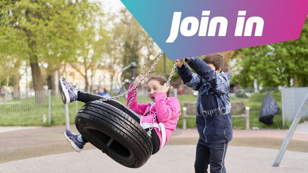 A girl plays on a tyre swing in a playground pushed by a boy, his face is hidden and she is laughing.