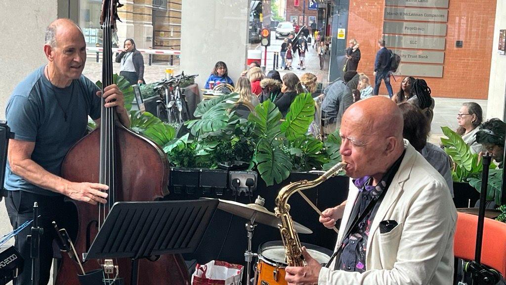 A man in a cream suit is playing a saxophone and a man in a dark blue T-shirt is playing a large string instrument. There are people milling around in the background.