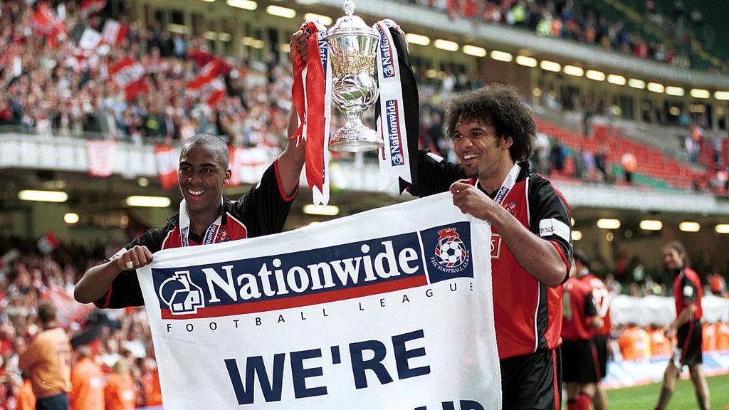 Darren Byfield, left, and Don Goodman, right, hold aloft the Division Two play-off winners' trophy and a sign reading 'We're Going Up'