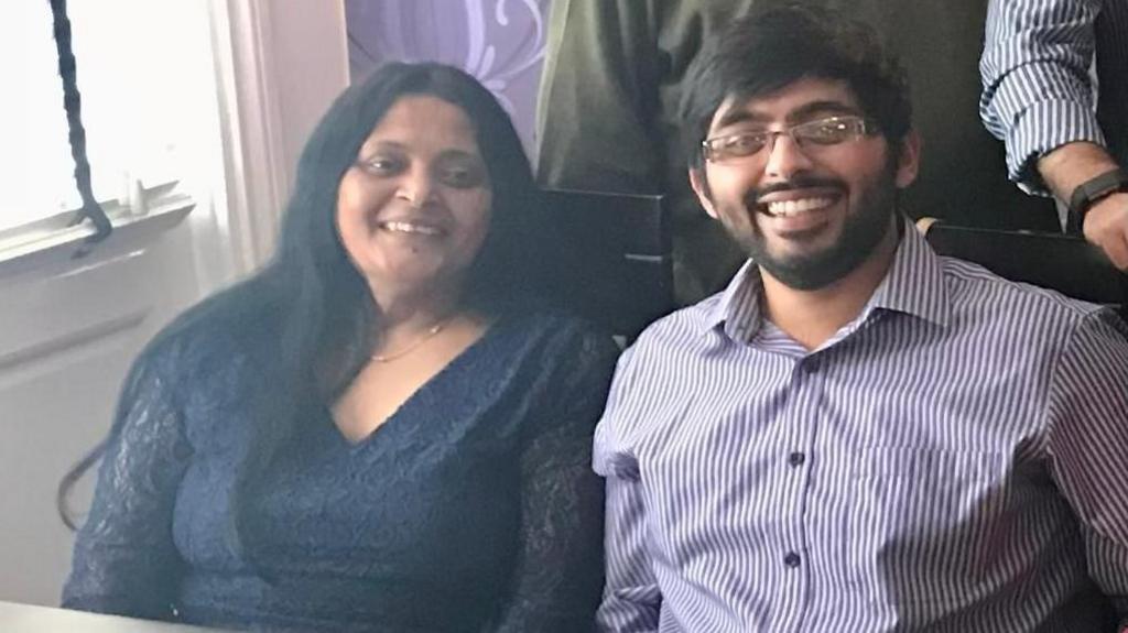 A woman with long dark hair and a blue v-neck top sits down next to her son, Arjun, who is wearing a striped white and purple shirt. He has dark hair and a beard and glasses.