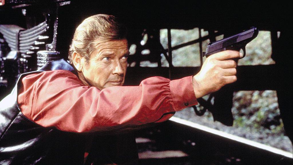 Actor Roger Moore on the set of Octopussy. He is wearing a red shirt and black waistcoat as he leans across a railway track holding a small black gun. 