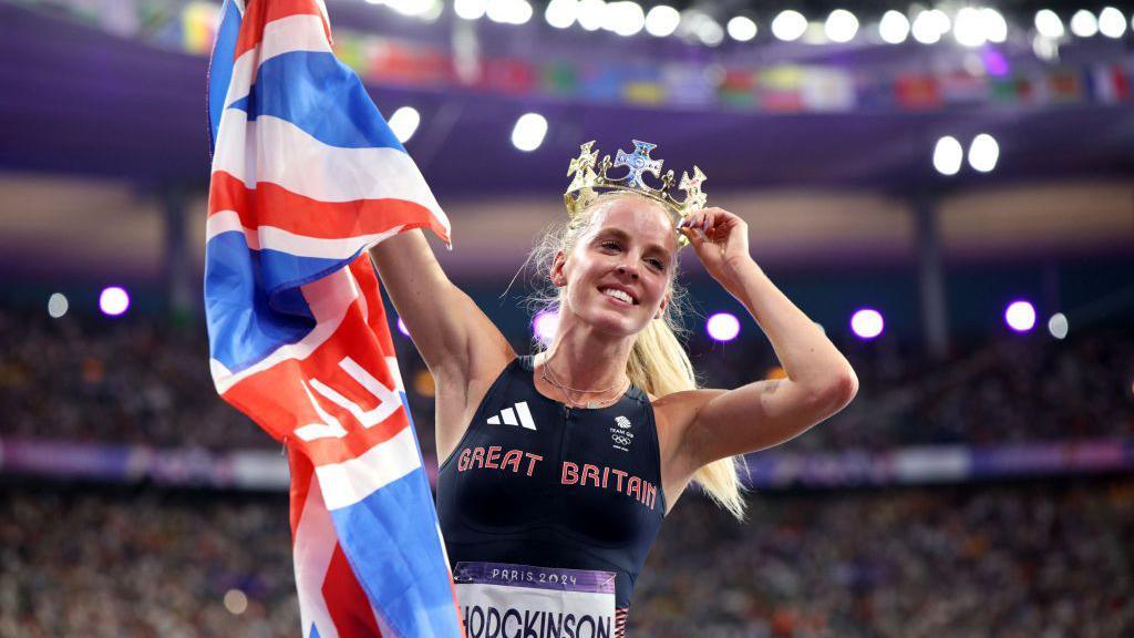 Keely Hodgkinson holds up the Union Jack with her right hand and places a crown on her head with her left after winning Olympic gold