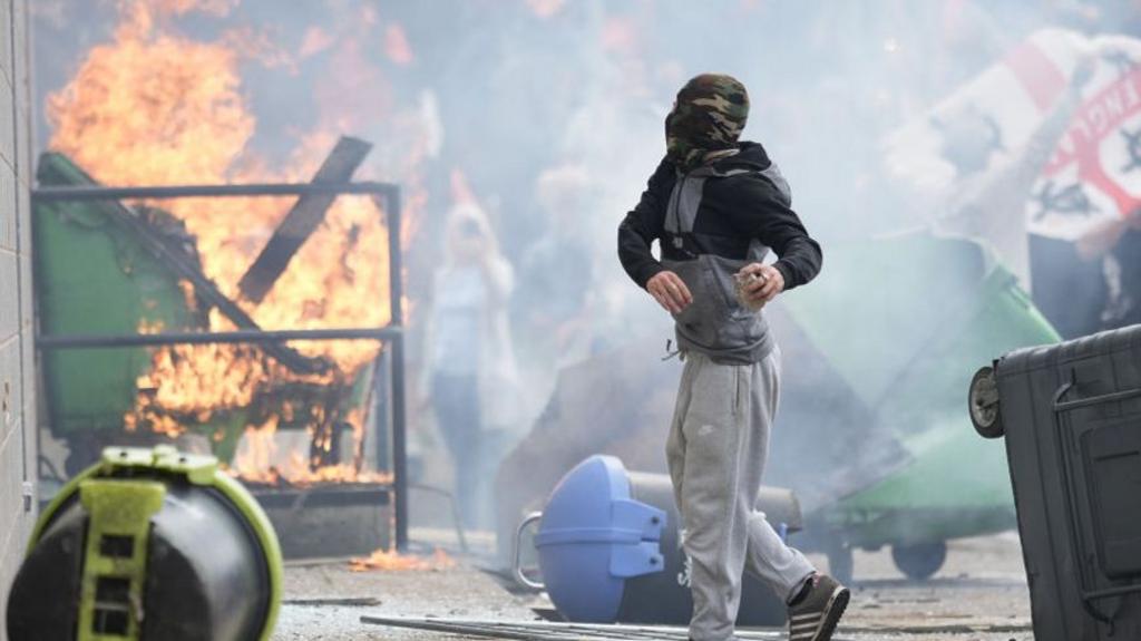 Masked person throws objects at hotel as large crowd is visible in background and a fire rages in the foreground