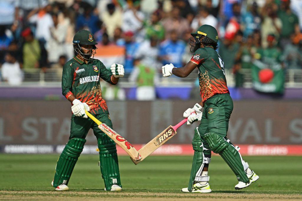 Two cricketers dressed in green prepare to fist bump while holding their cricket bats. There is a crowd of spectators behind them.