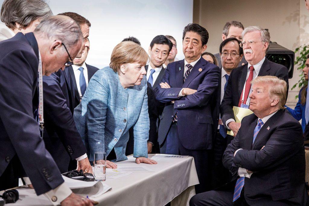 The then-German Chancellor Angela Merkel leans over a table to speak to a seated Donald Trump during a G7 summit in 2018 in Canada. They are surrounded by world leaders including Theresa May, UK prime minister, Emmanuel Macron, French president, Shinzo Abe, Japan prime minister, and John Bolton, US national security adviser.