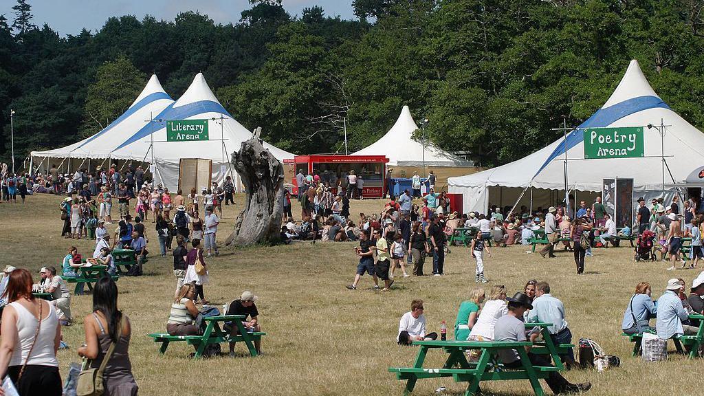 The Latitude Festival site in 2006 where three white tents had been erected in a grass field. Festivalgoers walk around the site where benches have also been placed.