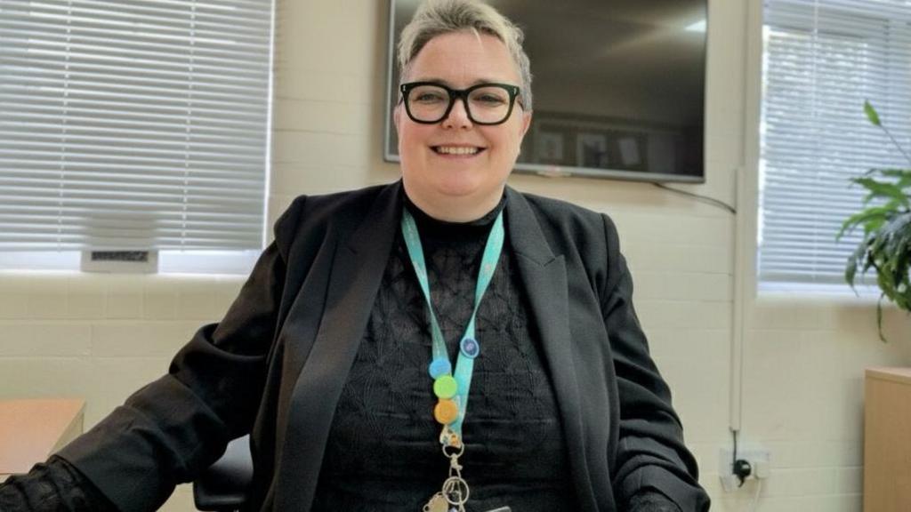 Kirsty Nichols-Mackay with short blonde hair, and black-framed glasses, wearing a black blazer and blouse, with a blue lanyard. She is sitting at a wooden desk and is smiling.