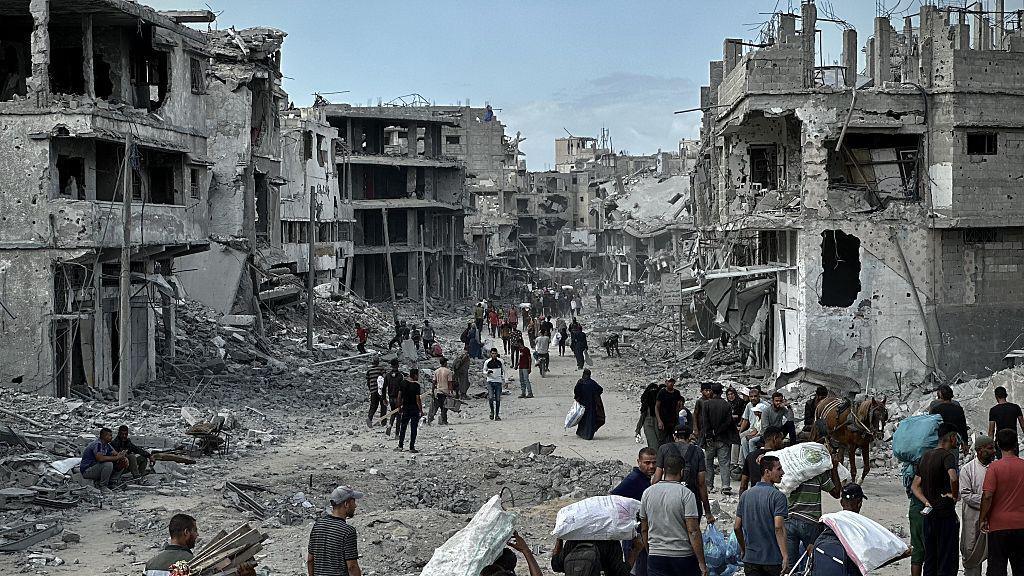 People walk with their belongings in the Khan Younis area of Gaza, surrounded by homes that are extremely damaged. Rubble is strewn on the street.