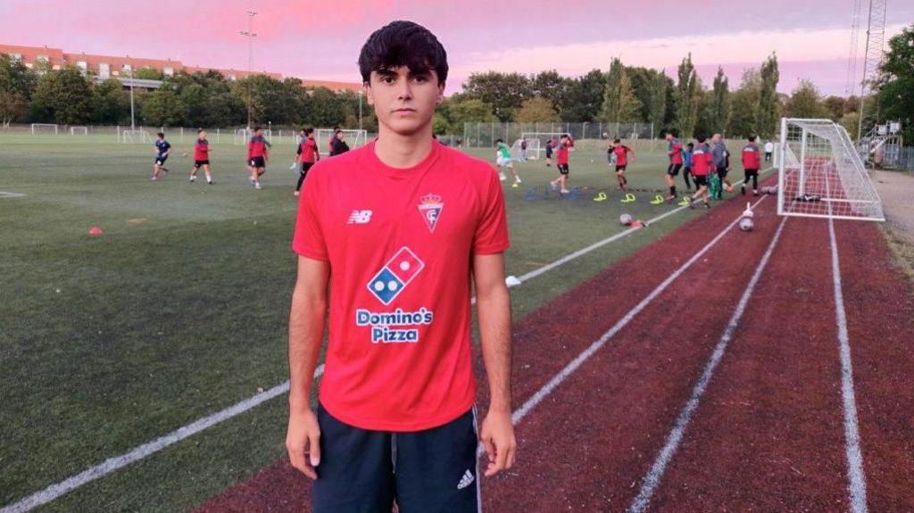 A young man in a red T-shirt and sports kit stands facing the camera with a stern face on the edge of a football pitch with a team playing in the background and a sunset sky.