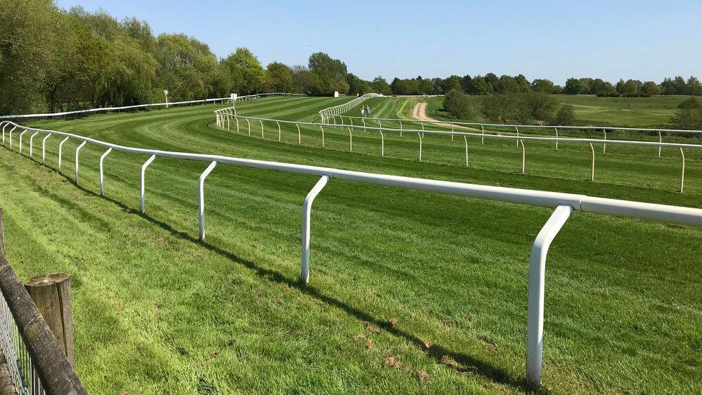 An empty grass racecourse with trees surrounding the perimeter and a white fence marking the track. 