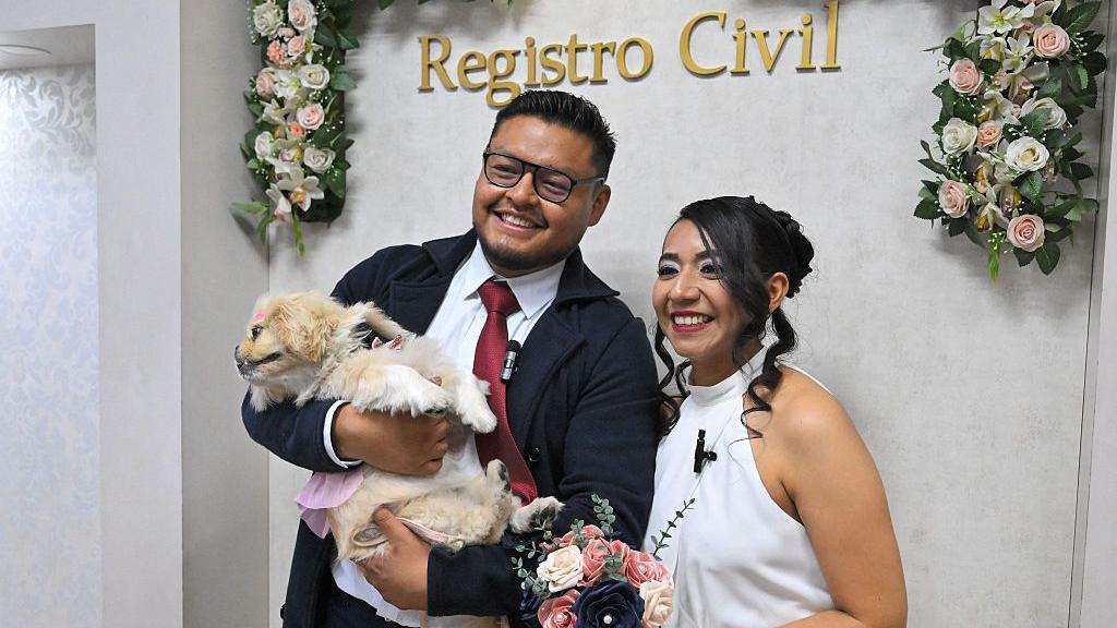 Andres and Diana pose with flowers, as Andres holding Luna who is dressed in a pink dress and bow.