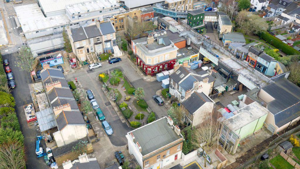 An aerial image of Albert Square. A small park can be seen in the middle of a square of terraced townhouses, with the famous red-fronted Queen Vic on the corner.