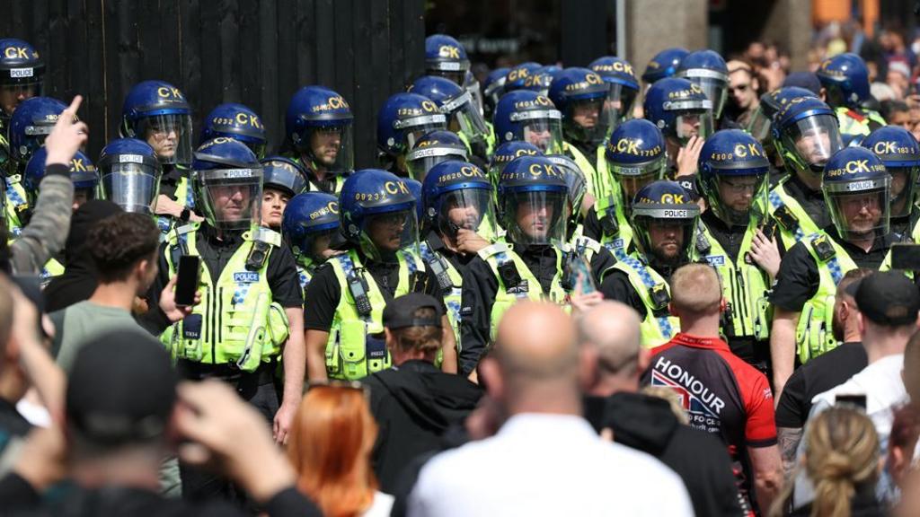 Police officers in riot gear stand guard as people participate in a protest in Manchester