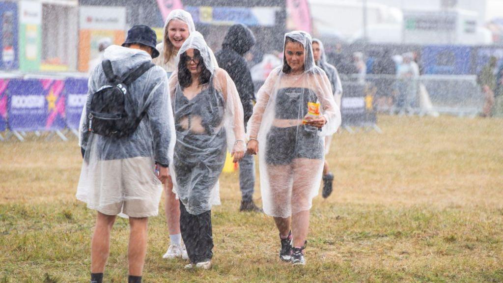 Four people wearing plastic ponchos shelter from the rain at Reading Festival.