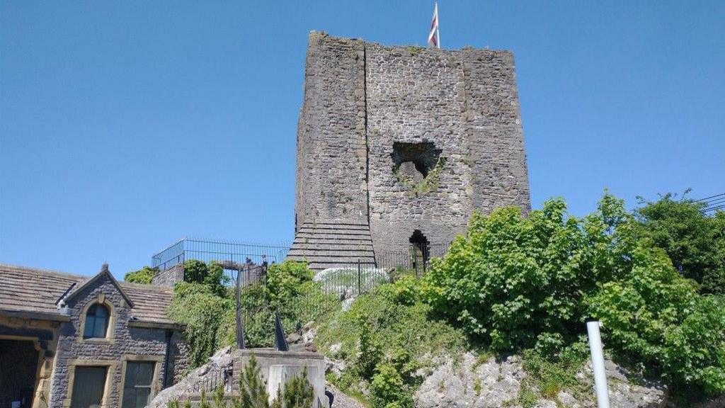 The stone keep of the castle can be seen at the top of a hill, with a flag on top. There are steps leading up to it, with a stone wall and stone building next to it, alongside shrubbery.