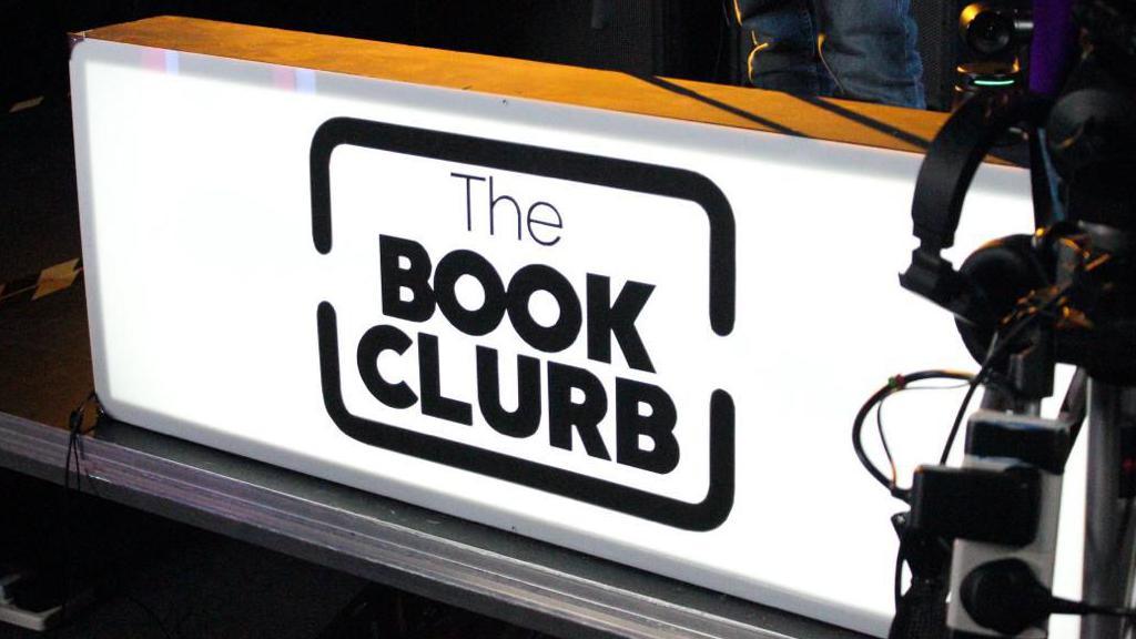 An illuminated sign that says The Book Clurb in back text on a white rectangle background.