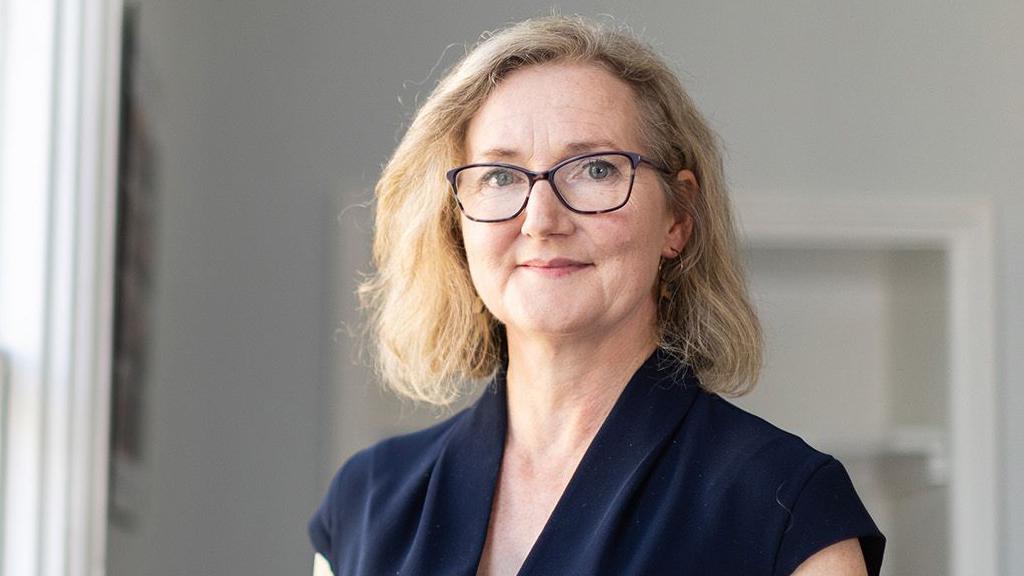 Photo of Rosie Phillips - CEO of Developing Health and Independence. She is looking at the camera, and is wearing a blue top, dark framed glasses and has blonde hair