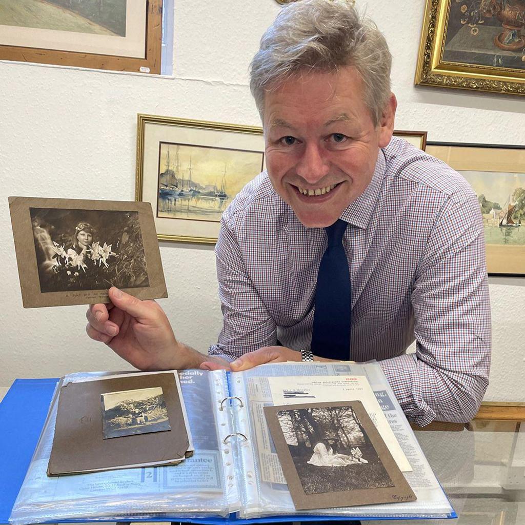 A man with grey hair, a purple and white square patterned shirt and a navy tie is holding a black and white photo of a girl with fairies in front of her. Behind him are paintings on a wall.
