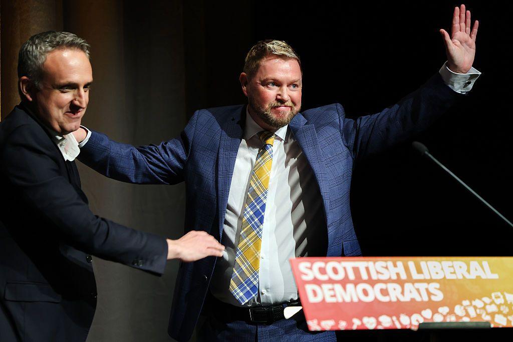 Jamie Greene MSP - a blond-haired man with a beard, wearing a dark suit with a jazzy tartan tie - waving triumphantly on stage at the Lib Dem conference, alongside grinning party leader Alex Cole-Hamilton