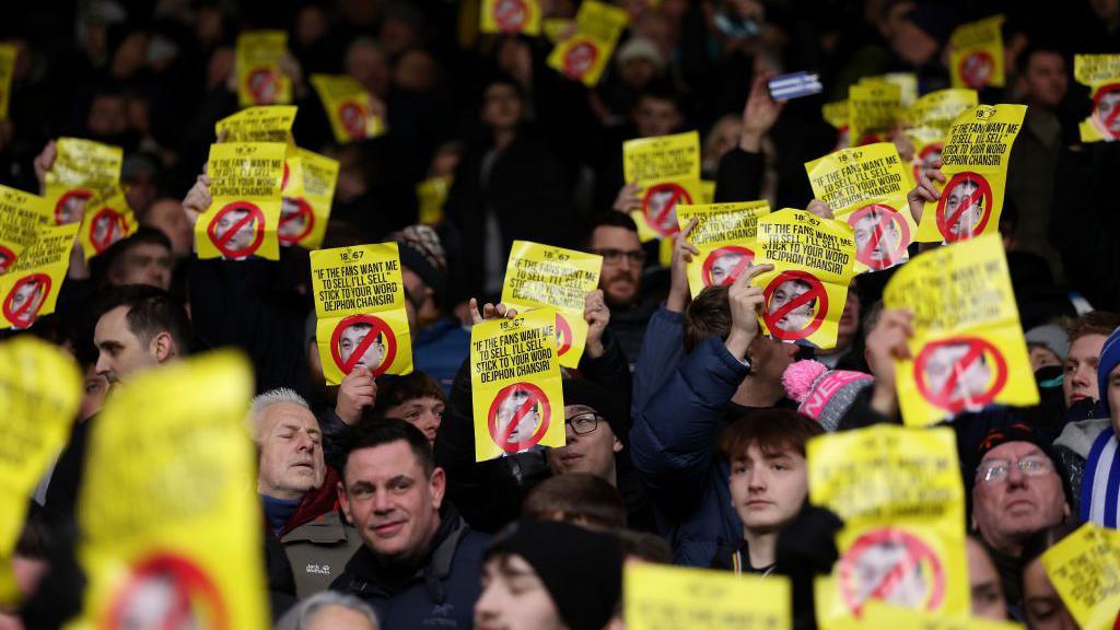 Sheffield Wednesday supporters in an away end hold up printed out signs bearing Dejphon Chansiri's face with a red cross, alongside a variety of slogans
