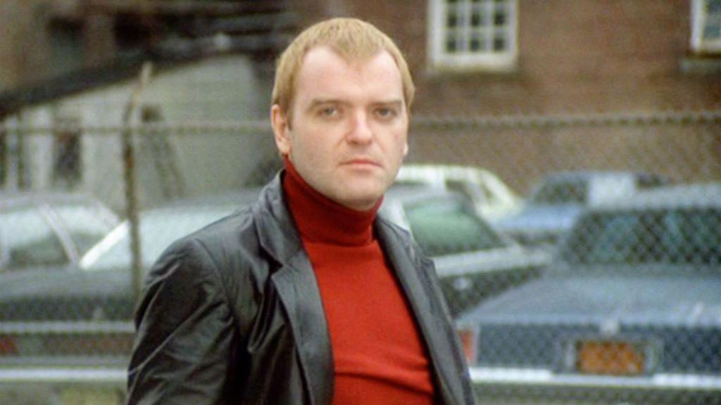 Angus MacInnes looking straight at the camera. He is wearing a red polo neck jumper under a black leather jacket and has fair hair.