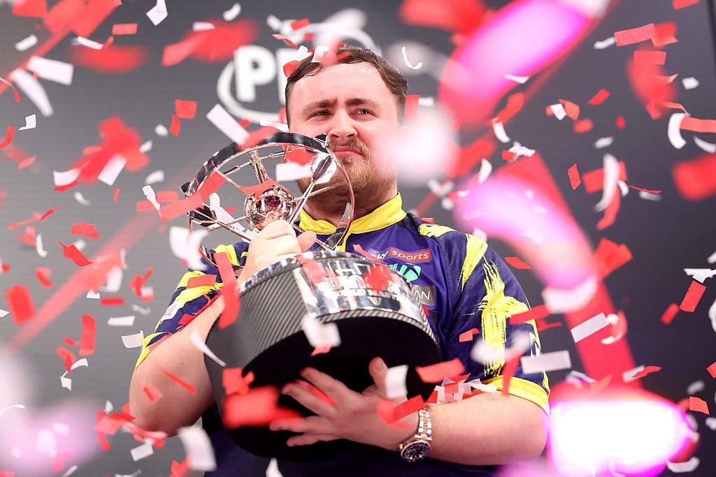 Darts player Luke Littler lifts the Winmau World Masters trophy as red and white confetti rains down, capturing a powerful moment of victory celebration.