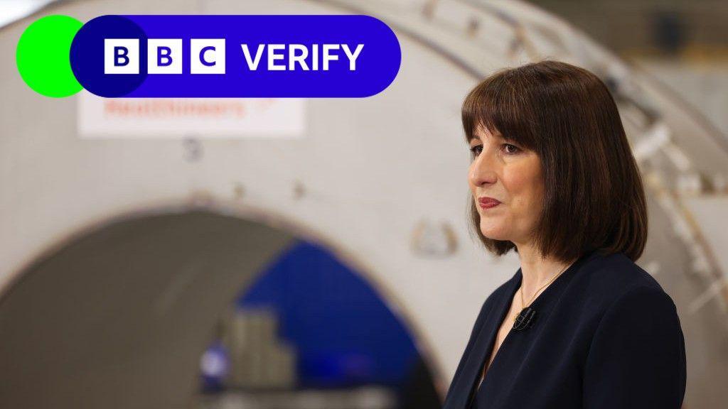 Rachel Reeves speaks at a news conference. She is wearing a dark jacket and is standing side on to the camera. In the background, a large machine can be seen. 